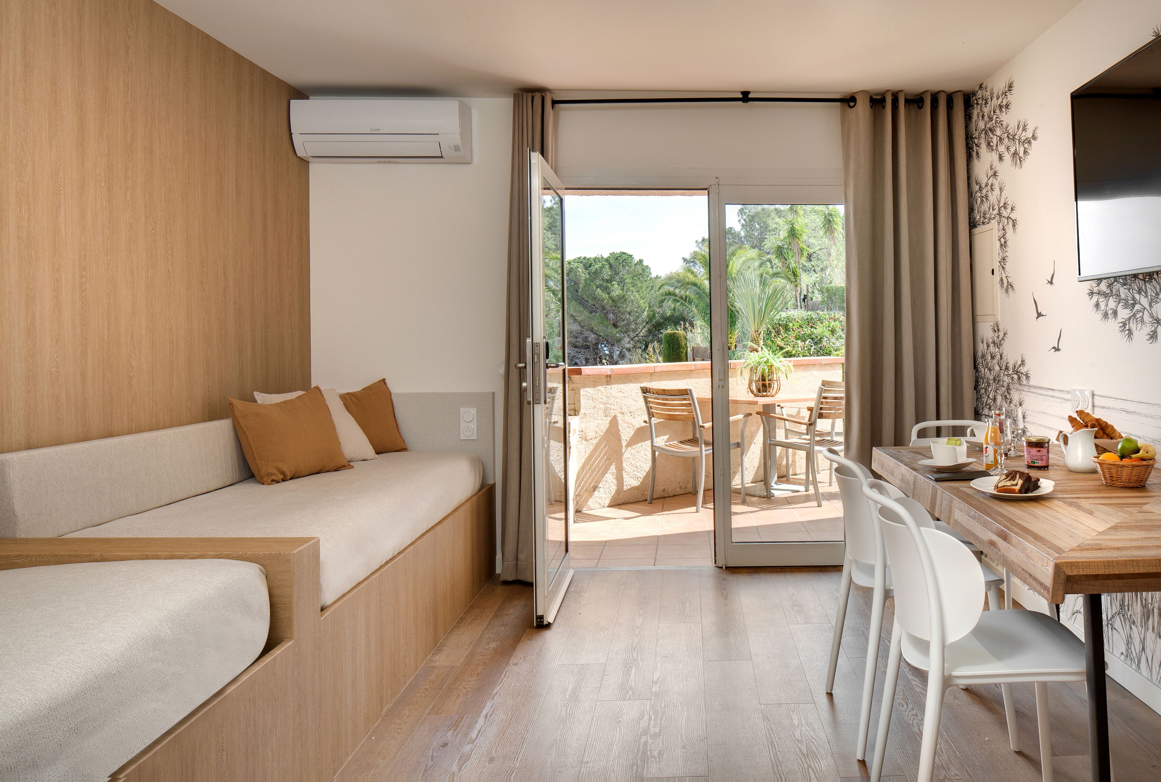 A modern hotel room with a sofa, dining table, chairs, and a balcony with outdoor seating.