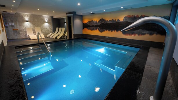 A modern indoor swimming pool with clear water, a mural of mountains, and a set of sun loungers. - Hotel Ariston (Pinzolo)