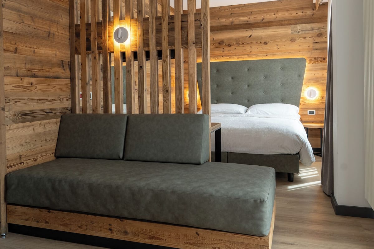 Un dormitorio moderno con separador de listones de madera, un sofá gris, una cama con cabecera gris y una mesita de noche con lámpara.