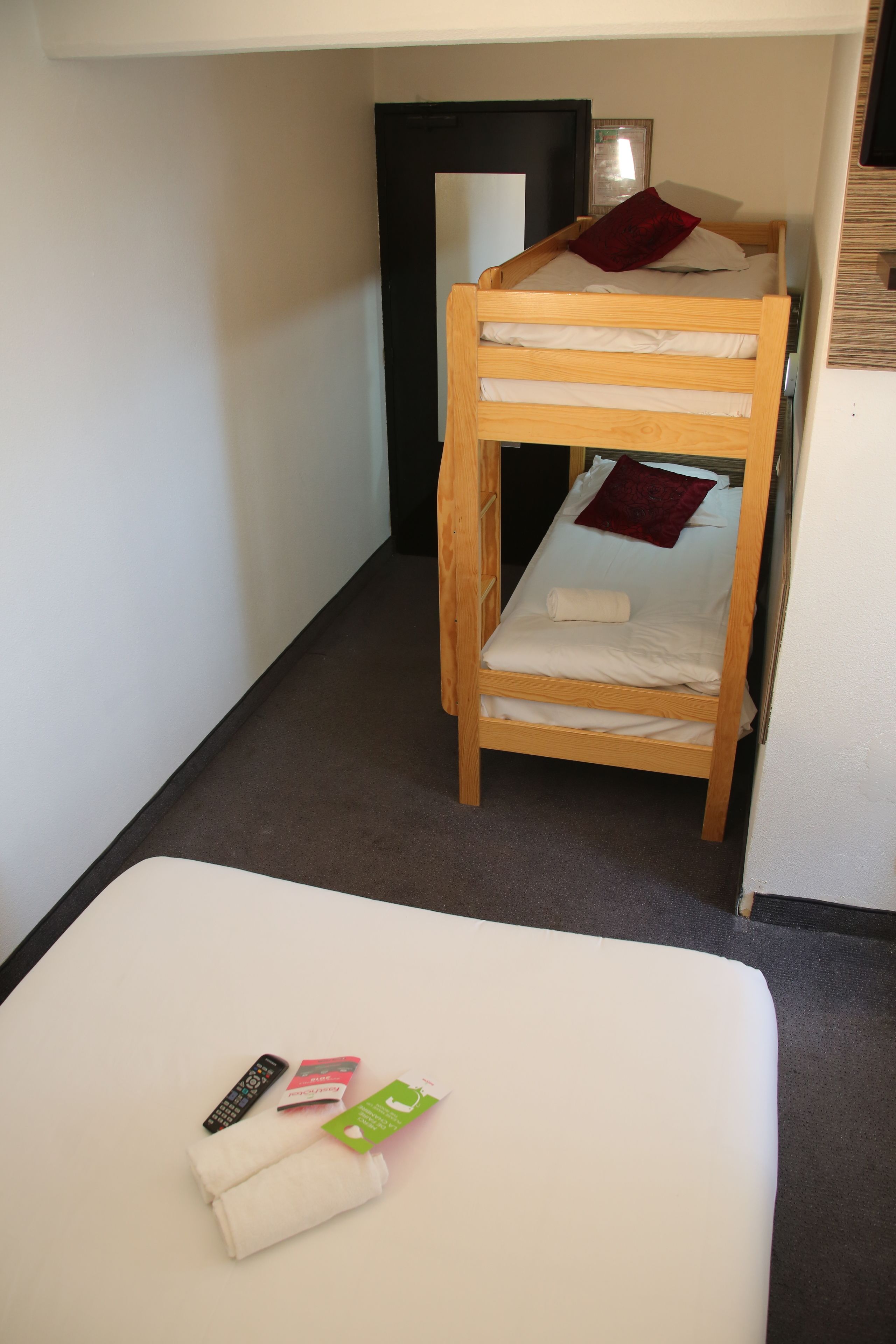 Een stapelbed met een houten frame, een witte matras en een rood kussen. Op een witte tafel liggen een afstandsbediening, een boek en een groene kaart.