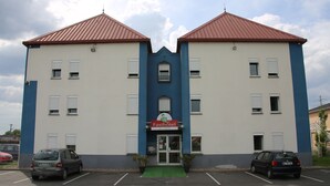 Un bâtiment à deux étages avec une entrée centrale et une enseigne indiquant « Fasthotel ». Deux voitures sont garées devant.