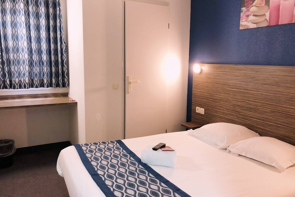 Une chambre d’hôtel avec un lit, une table de chevet, une commode, une fenêtre avec des rideaux à motifs et un tableau accroché au mur.