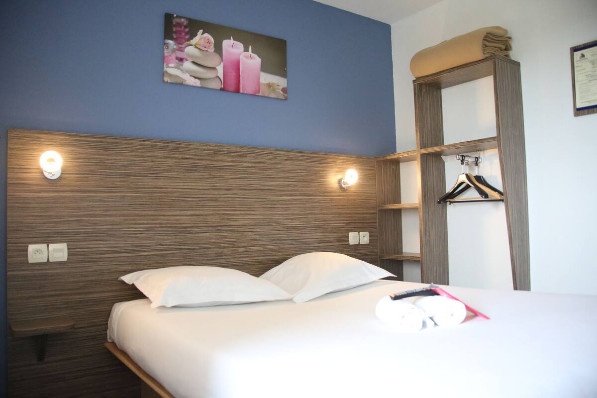 Une chambre d’hôtel avec une tête de lit en bois, un lit recouvert de draps blancs, une lampe fixée au mur, un porte-manteau et un tableau encadré au-dessus du lit.