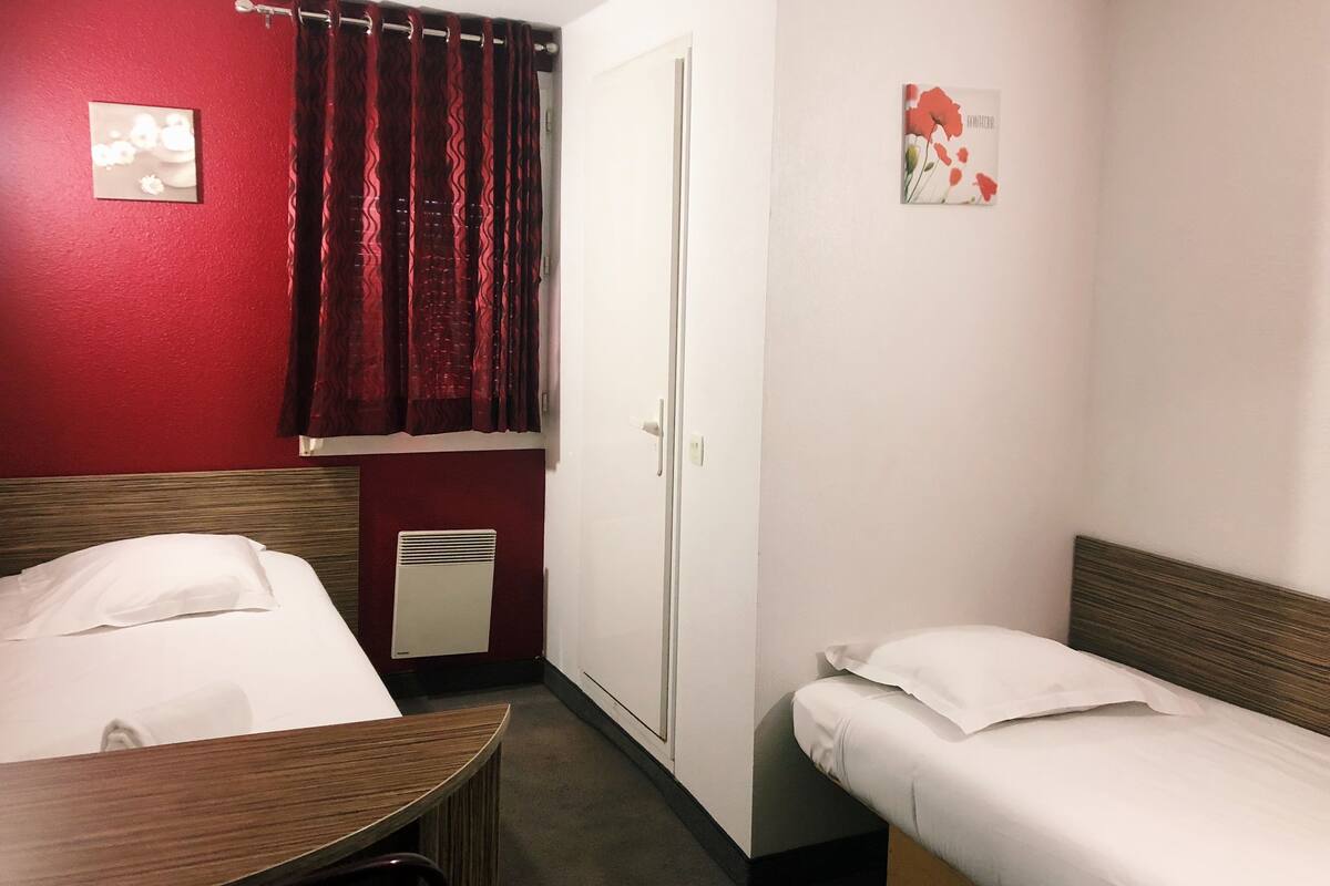Une chambre d’hôtel avec deux lits simples, une table en bois, un mur rouge, une porte blanche et un petit radiateur.