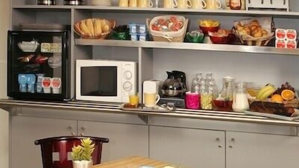 Une cuisine compacte équipée d’un four à micro-ondes, d’une machine à café et de divers produits alimentaires rangés sur des étagères.