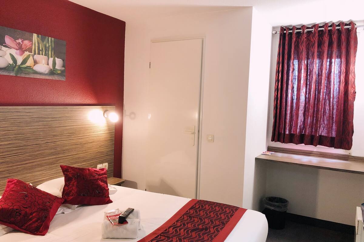 Une chambre d’hôtel avec un lit, des oreillers rouges, un tableau au mur, une porte et une fenêtre avec des rideaux.