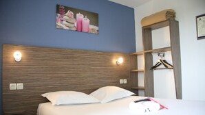 Une chambre d’hôtel avec une tête de lit en bois, un lit avec du linge de lit blanc, une lampe fixée au mur, un tableau encadré et un porte-manteau.