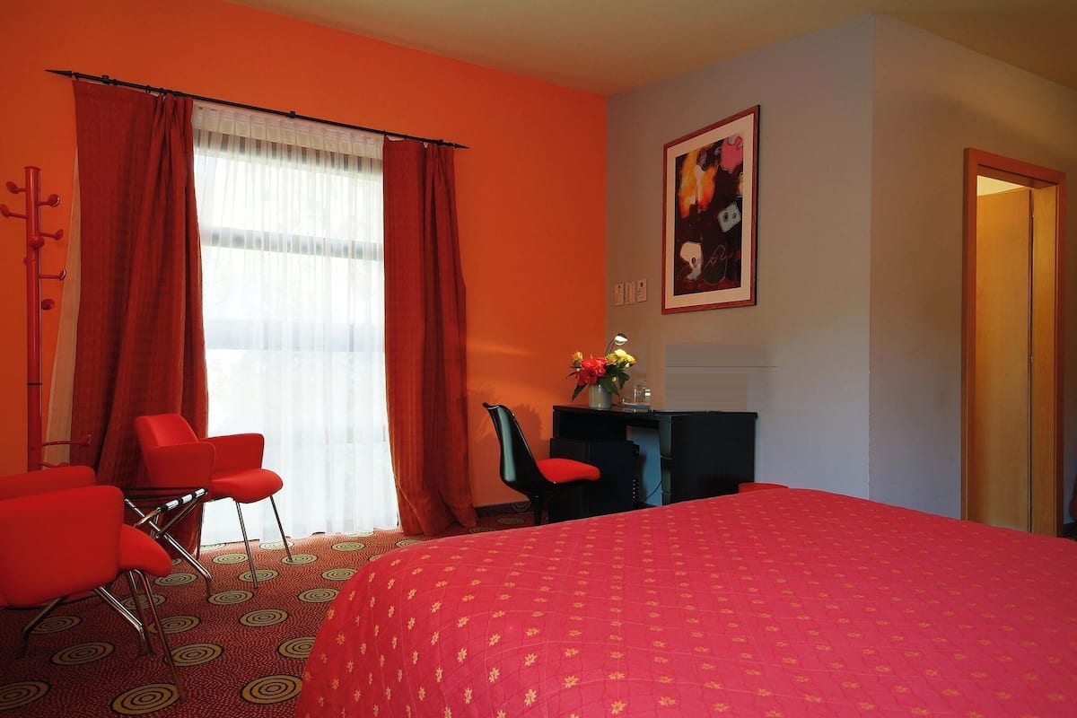 A hotel room with a large bed, red chairs, a desk with a vase of flowers, and a painting on the wall.