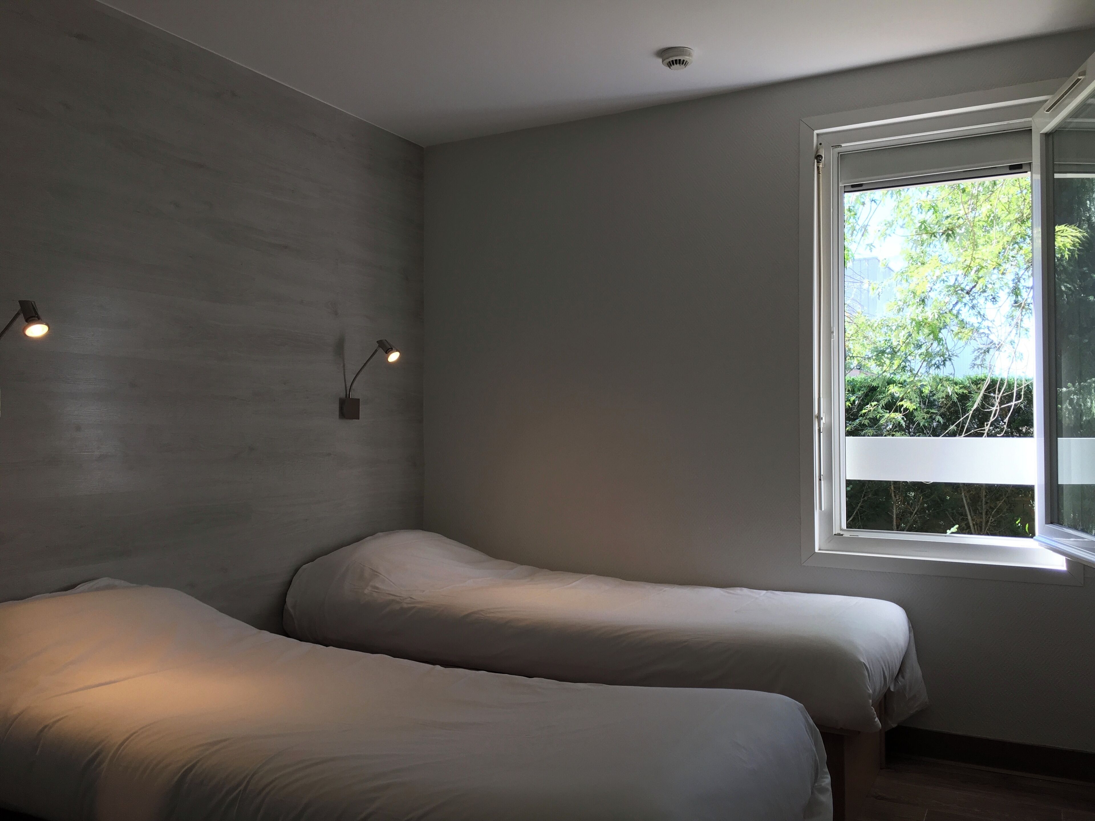 Une chambre moderne avec deux lits, une grande fenêtre donnant sur un paysage verdoyant et un luminaire fixé au mur.