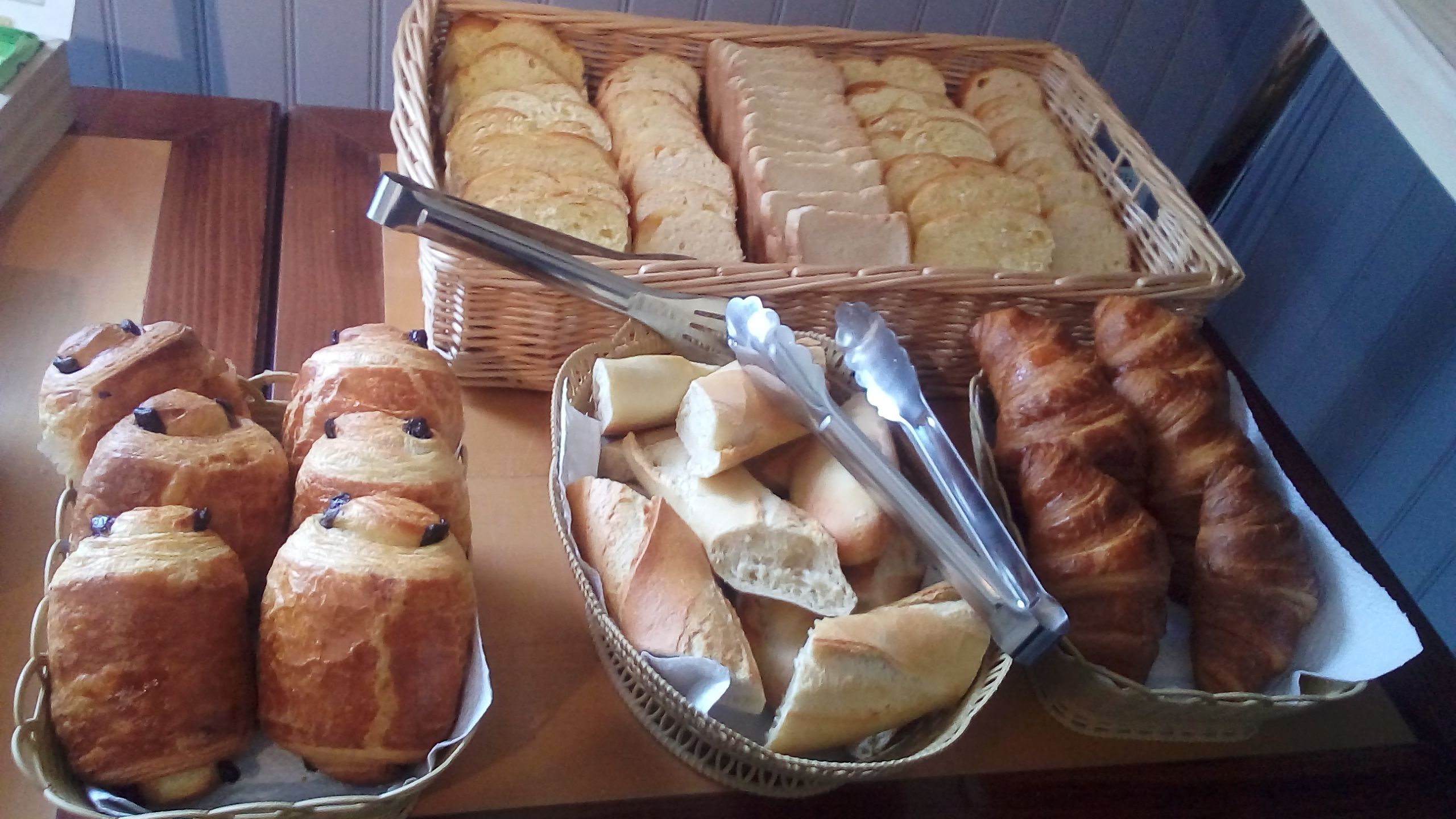 Pöydällä on erilaisia leipiä ja leivonnaisia, kuten croissantteja ja patonkeja, jotka on aseteltu koreihin ja tarjottimelle.