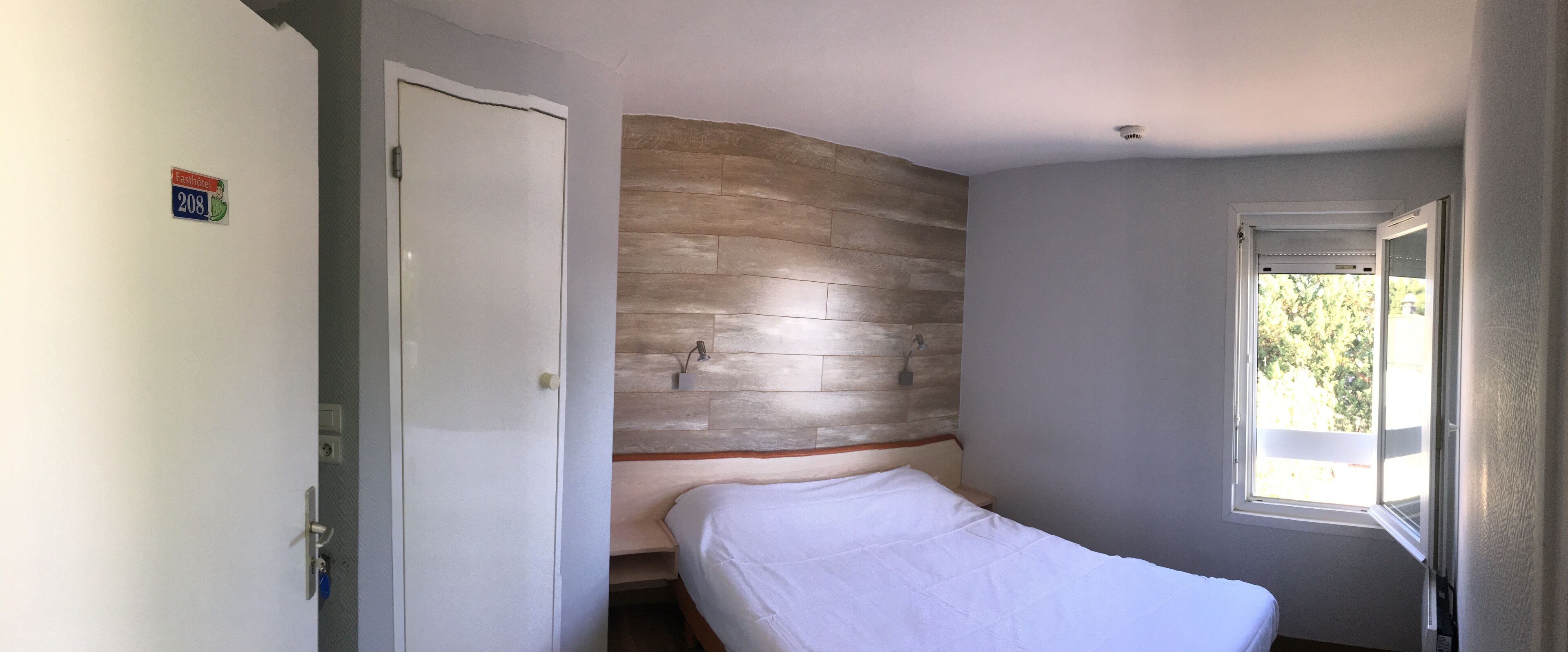 A bedroom with a wooden headboard, a bed with a white mattress, a door with a number plate, and a window with a view of greenery.