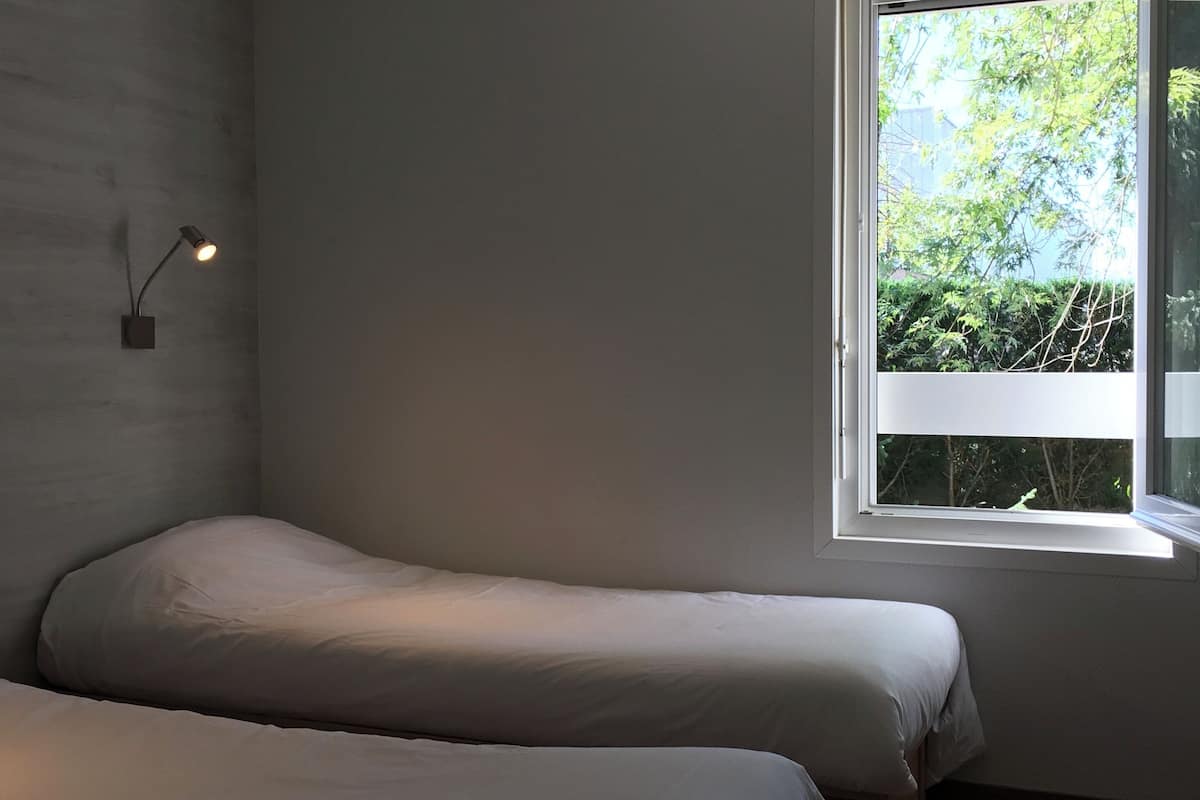 A bedroom with two single beds, a window with a view of greenery, and a wall-mounted light fixture.