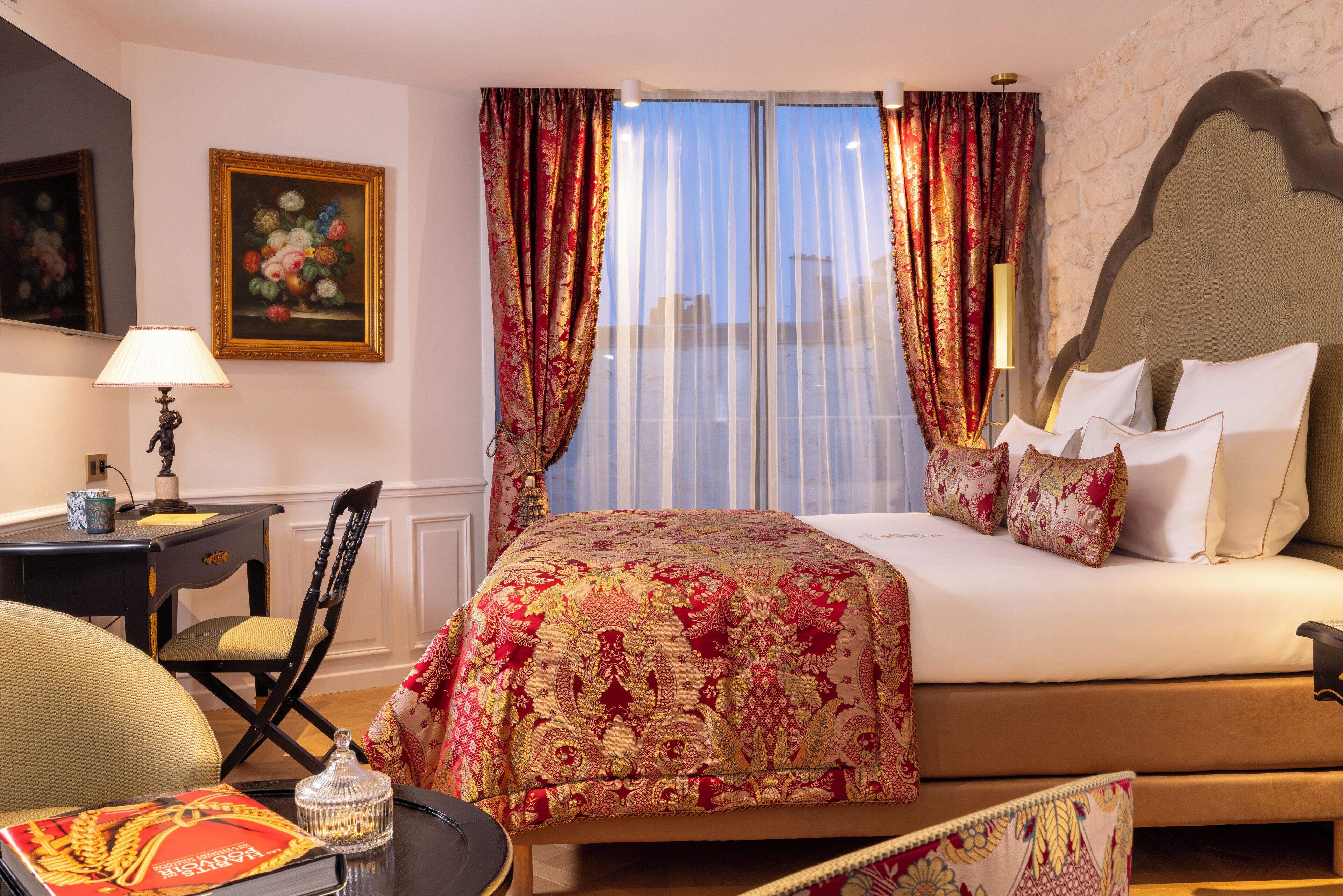 A hotel room with a large bed, a desk with a lamp, a chair, and a view of the city through sheer curtains.
