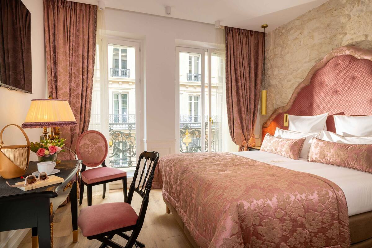 A hotel room with a large bed, a desk with a lamp, a chair, and a view of buildings through the windows.