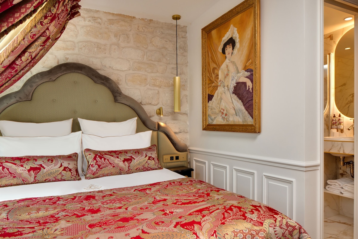 A bedroom with a large bed, ornate pillows, a classic headboard, a painting, and a bathroom visible through an open door.