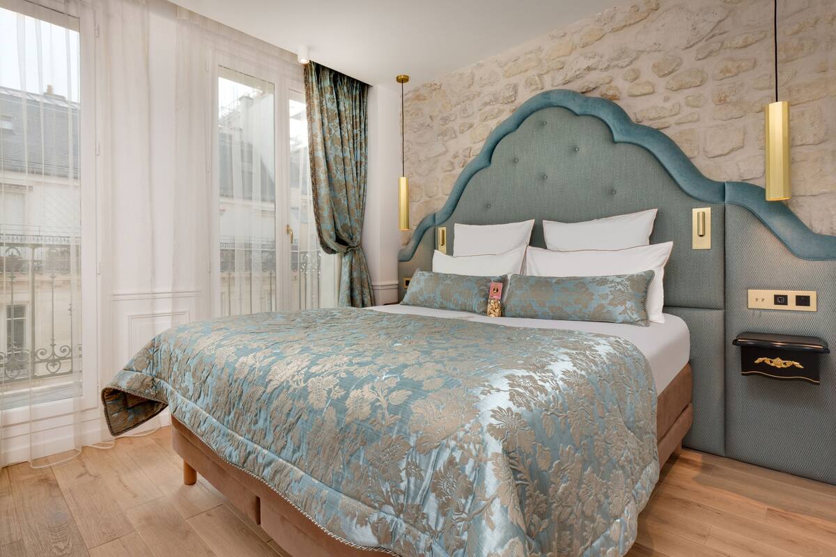 A bedroom with a bed, a stone wall, a window with curtains, and a nightstand with a lamp.