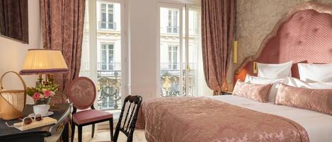 A hotel room with a large bed, a desk with a lamp, a chair, and a view of the city through large windows.