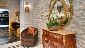 A lobby with a stone wall, a large ornate mirror, a brown armchair, and a wooden cabinet with a plant. - Hôtel Kleber Paris & Spa (Paris)