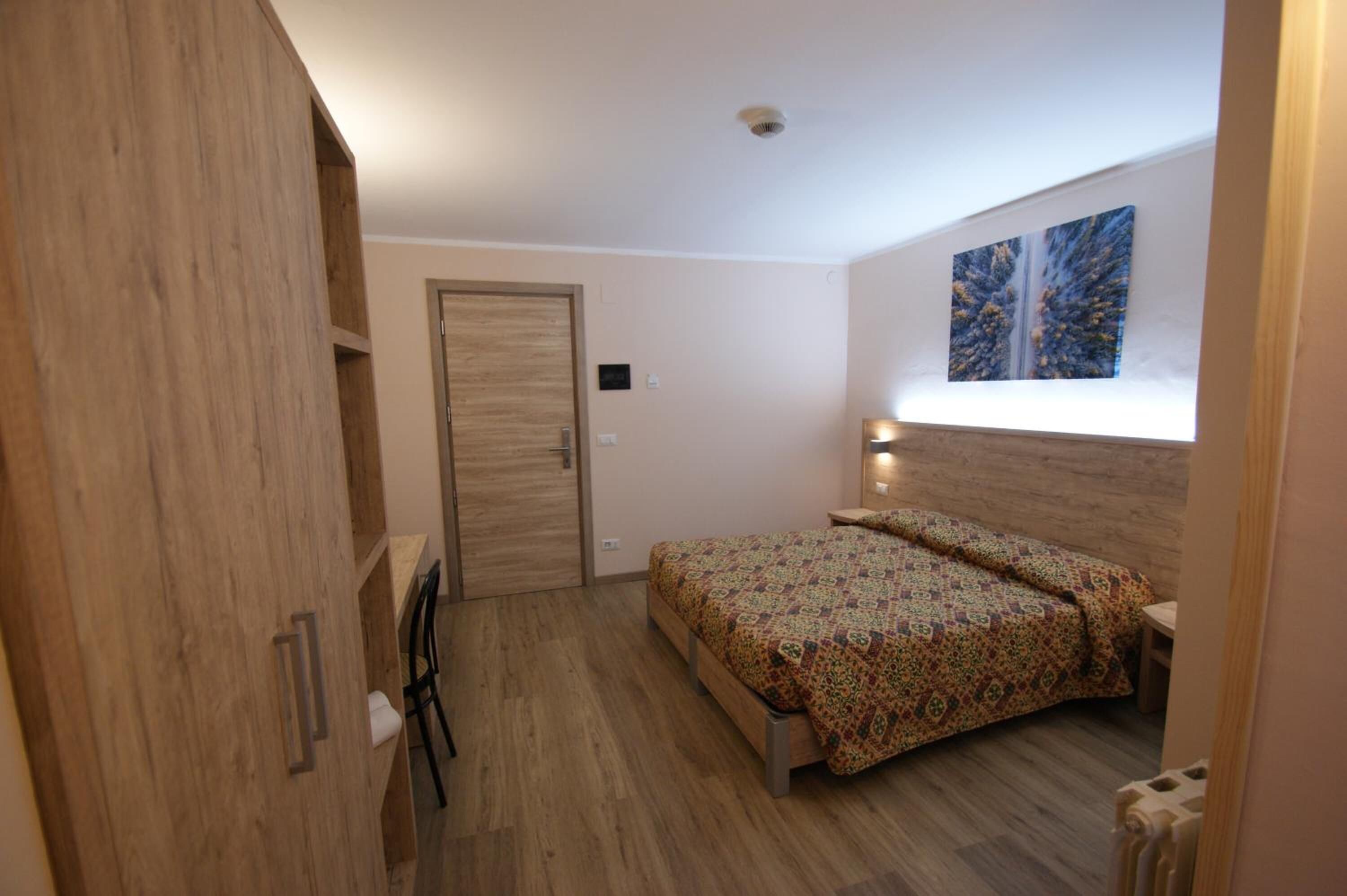 Ein Schlafzimmer mit einem Holzschrank, einem Bett mit gemusterten Textilien, einem Nachttisch mit einer Lampe und einem Fenster mit einem Kunstwerk.