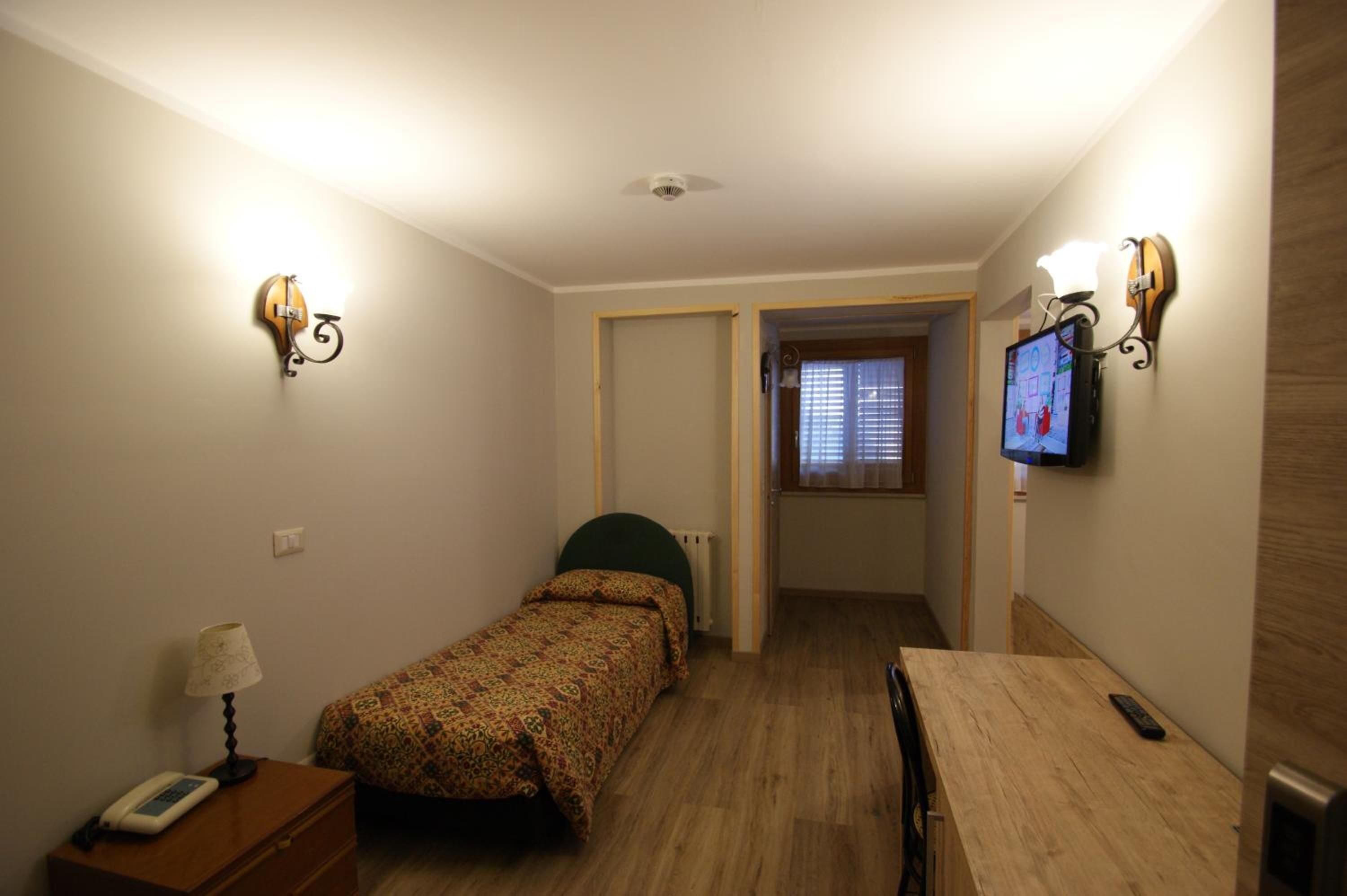 Una stanza piccola e stretta con un letto, un tavolo, una sedia, una lampada, un telefono e una televisione fissata al muro.