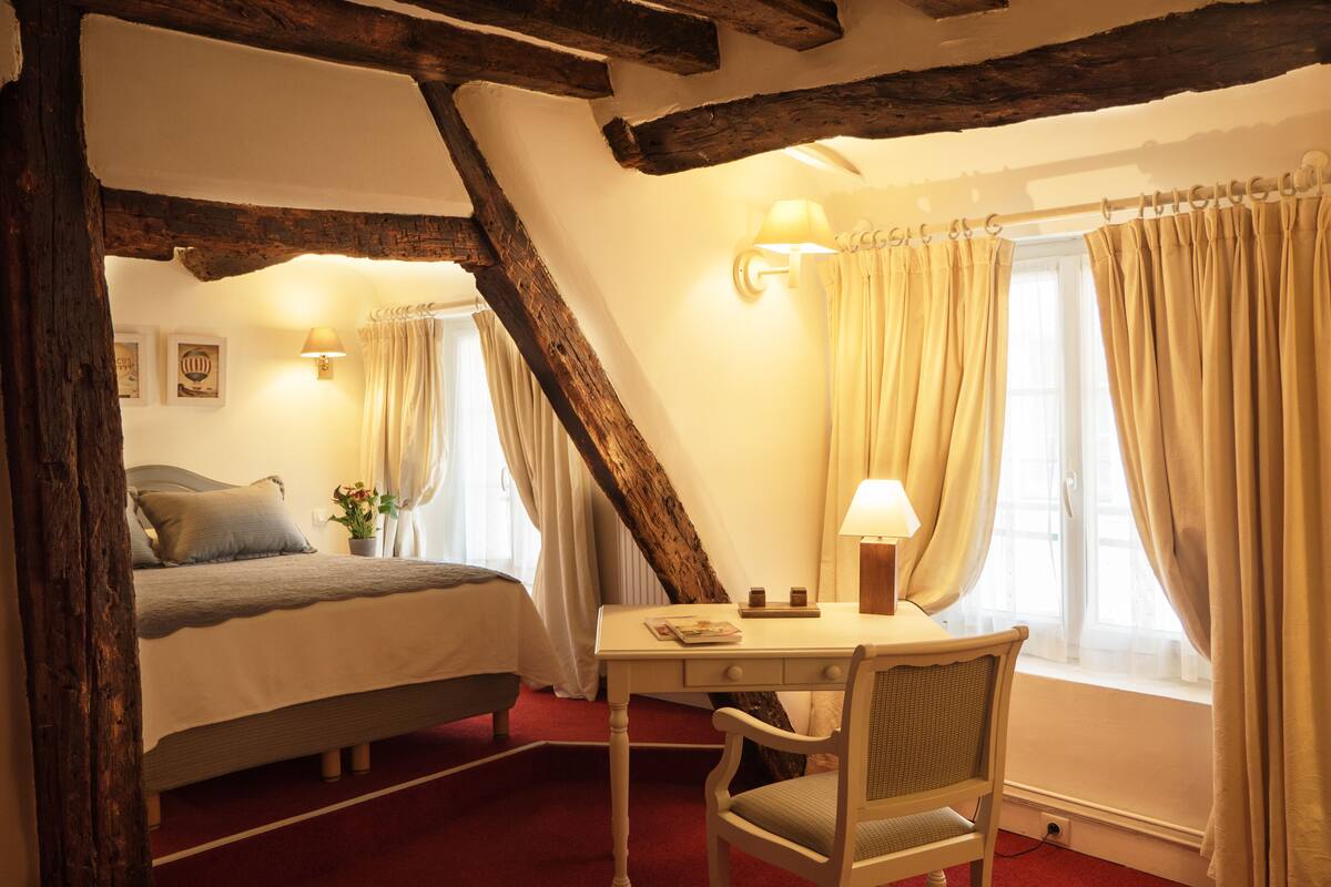 Ein Schlafzimmer mit einem Bett, einem Schreibtisch und einem Stuhl. Es gibt ein Fenster mit Vorhängen, eine Lampe und eine Wandleuchte.