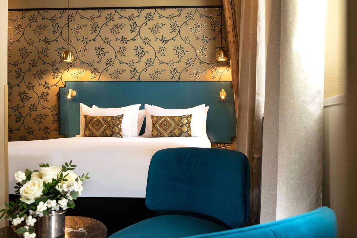 Ein Hotelzimmer mit einem Bett, einem blauen Sessel, einer Blumenvase und einem gemusterten Wandteppich.
