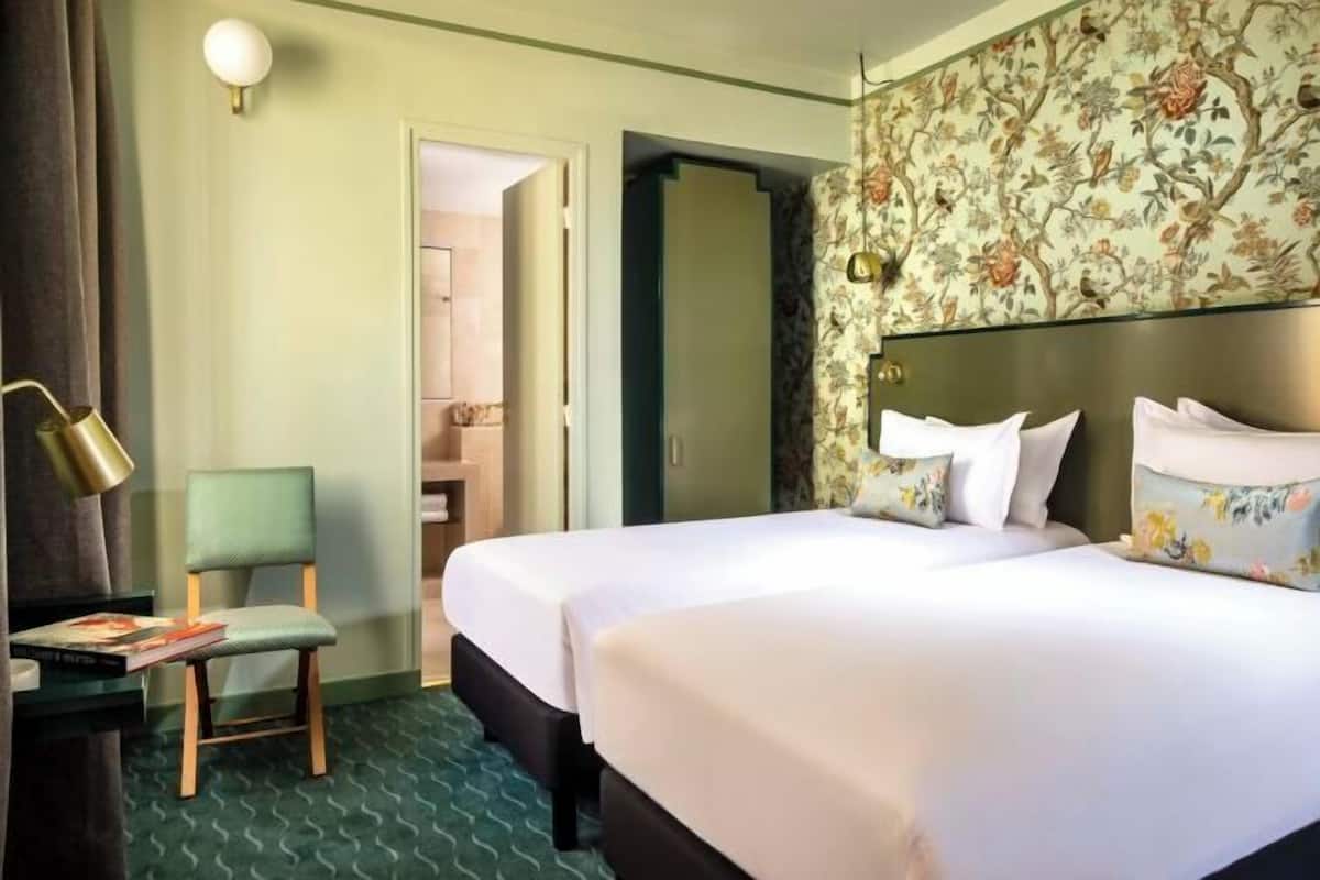 Ein Hotelzimmer mit zwei Betten, einem Sessel, einem Schreibtisch mit Lampe und Blumenmuster-Tapete.