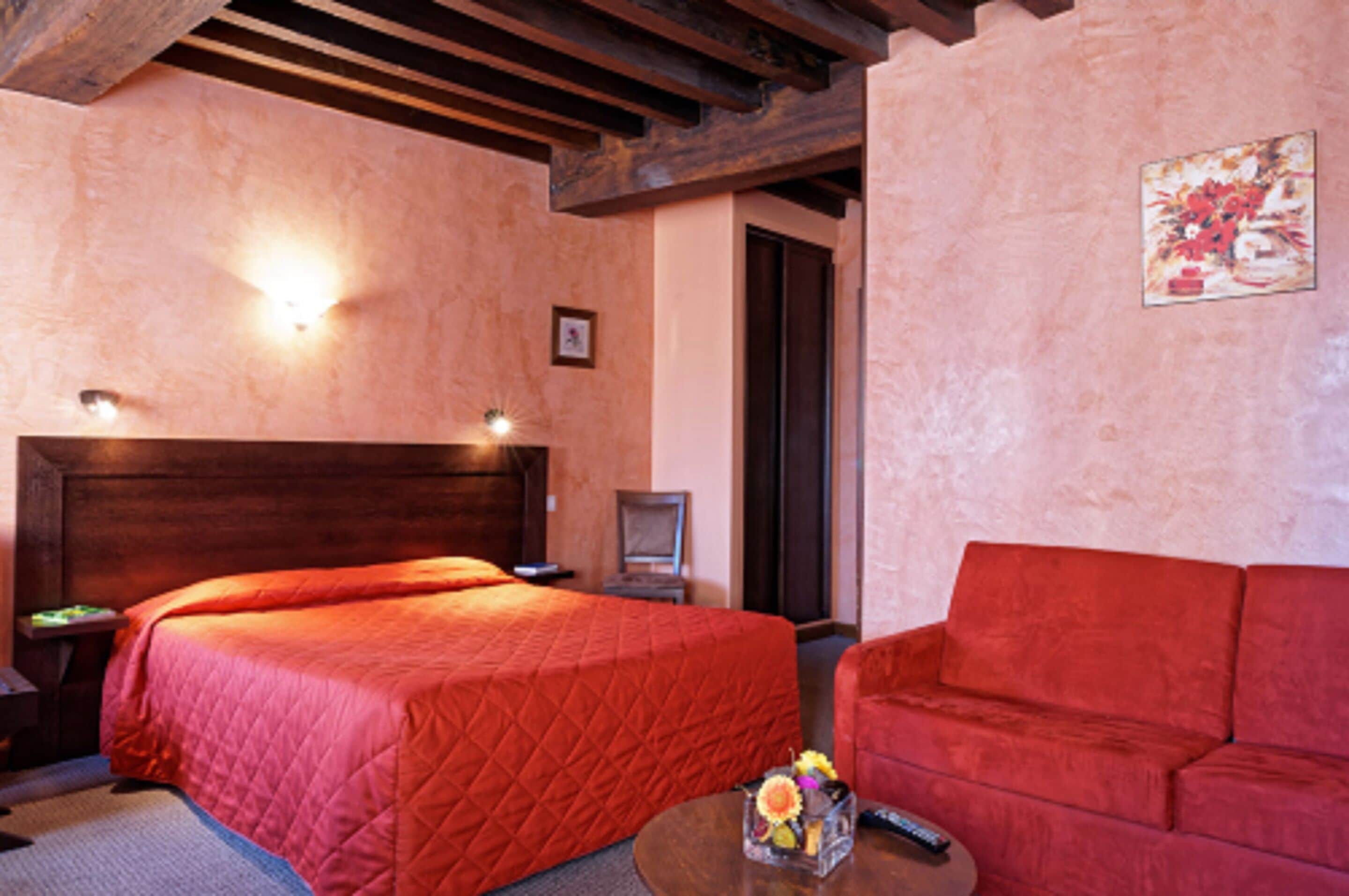 Een hotelkamer met een groot bed, een rode bank, een klein tafeltje met een bloevas en een schilderij aan de muur.