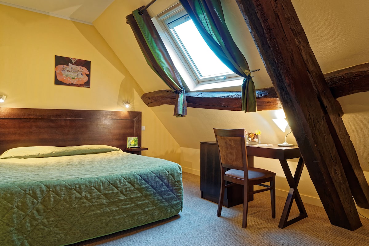 A bedroom with a bed, a desk, and a chair. There is a window with curtains, a painting on the wall, and a lamp on the desk.