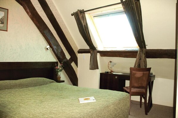 A room with a bed, a desk, and a chair. There is a window with curtains, a picture on the wall, and a small table with a plant.