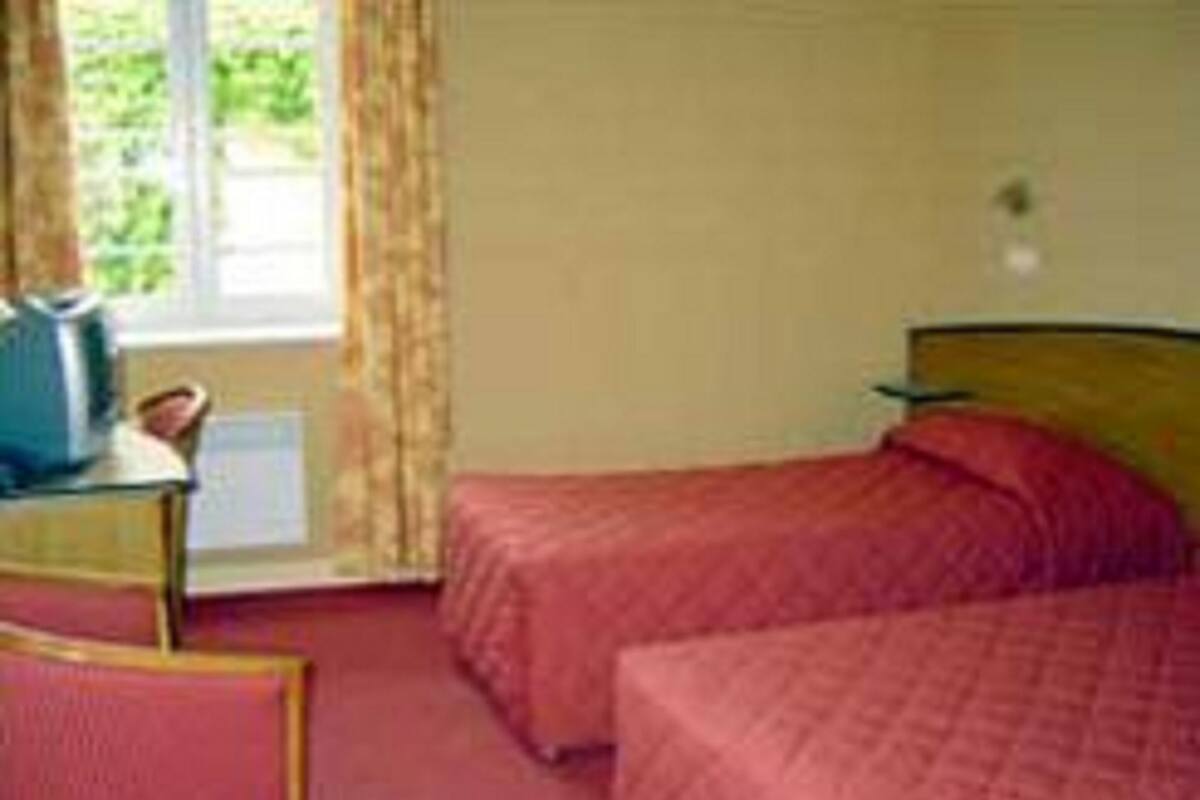 A hotel room with two beds, a small table with a chair, a window with curtains, and a view of greenery outside.