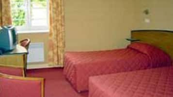 A hotel room with two beds, a small table with a chair, a window with curtains, and a view of greenery outside.