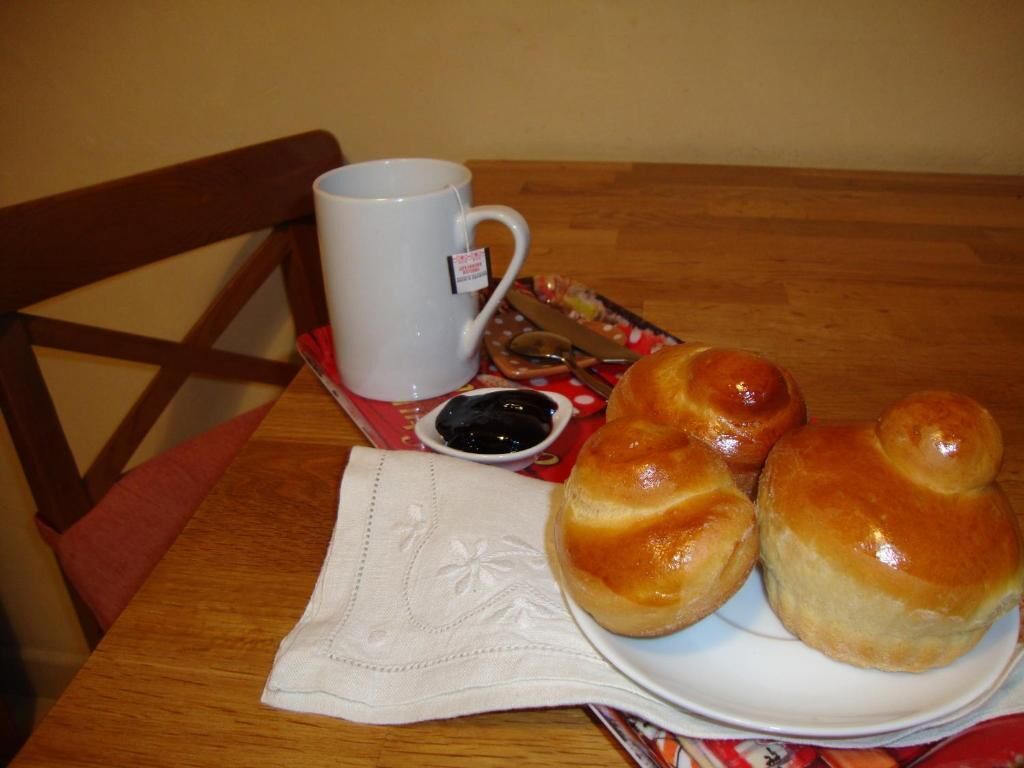 A wooden table with a white mug, a plate of bread rolls, and a small bowl of jam.