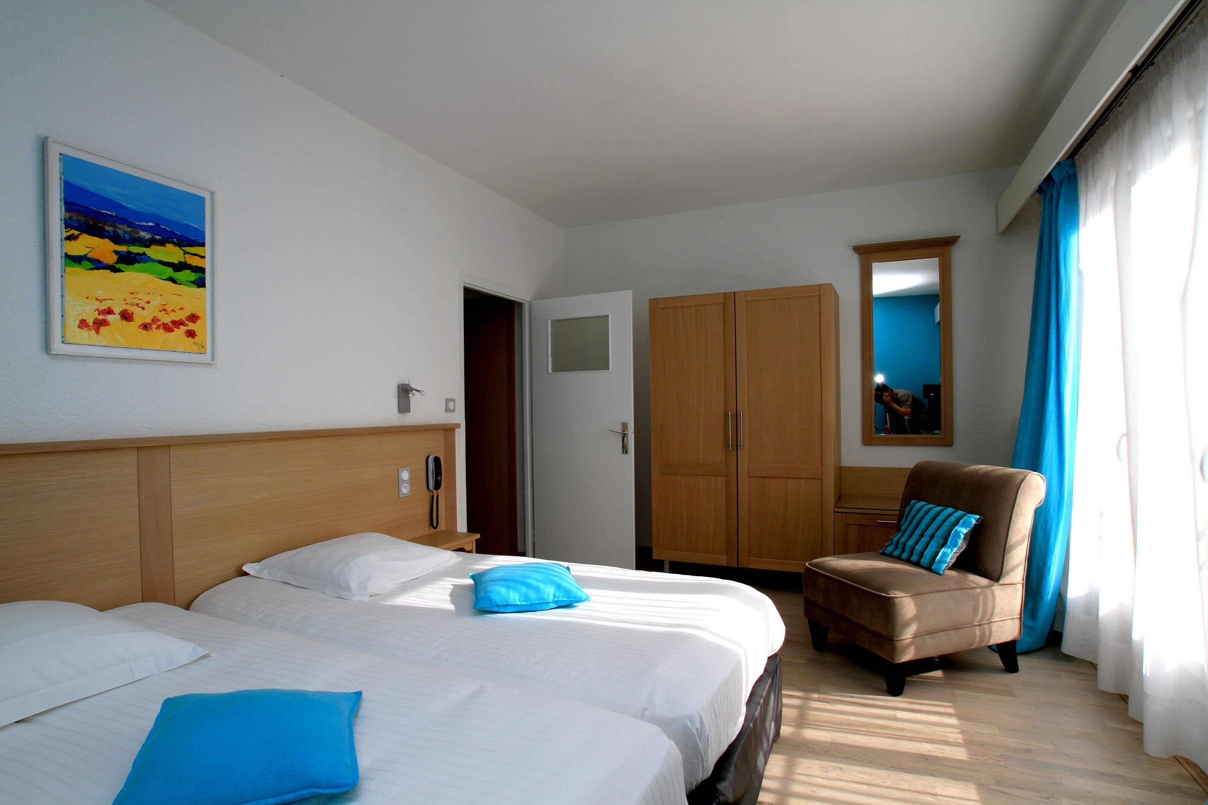 Habitación de hotel con dos camas, un armario de madera, un sillón azul y un cuadro en la pared.