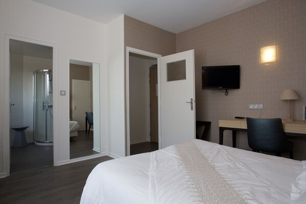 A hotel room with a bed, a desk, a chair, a TV, and a shower area. - LOGIS Beau-Site (Luxeuil-les-Bains)
