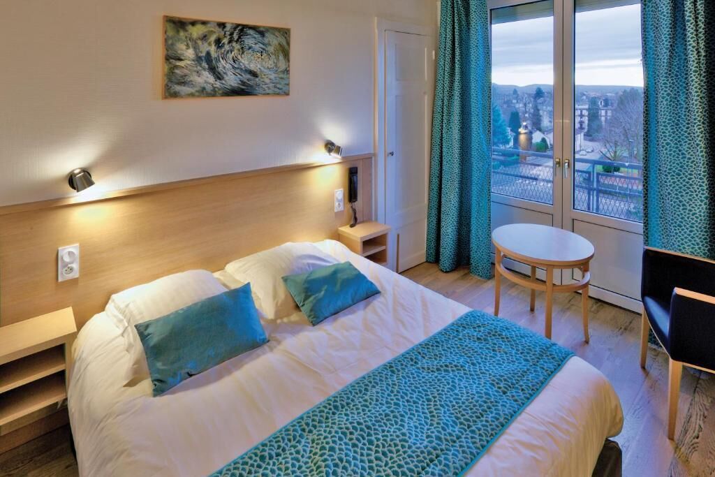 A hotel room with a bed, bedside tables, a small table, a chair, and a view through a window.