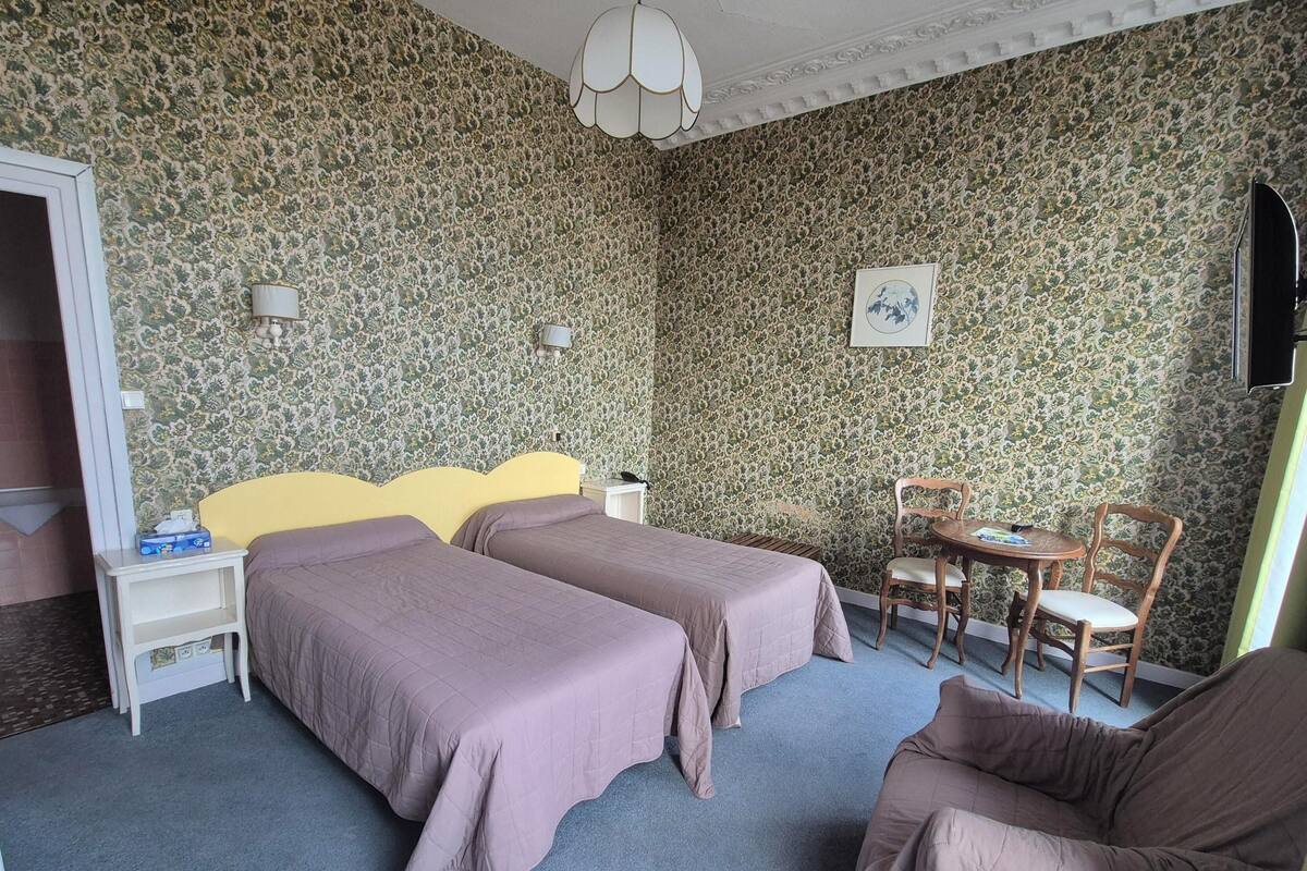 A hotel room with two beds, a small table with a chair, a lamp, and a painting on the wall.
