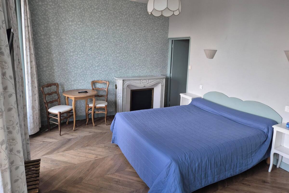 A bedroom with a large bed, a small round table with chairs, a fireplace, and a hanging light fixture.
