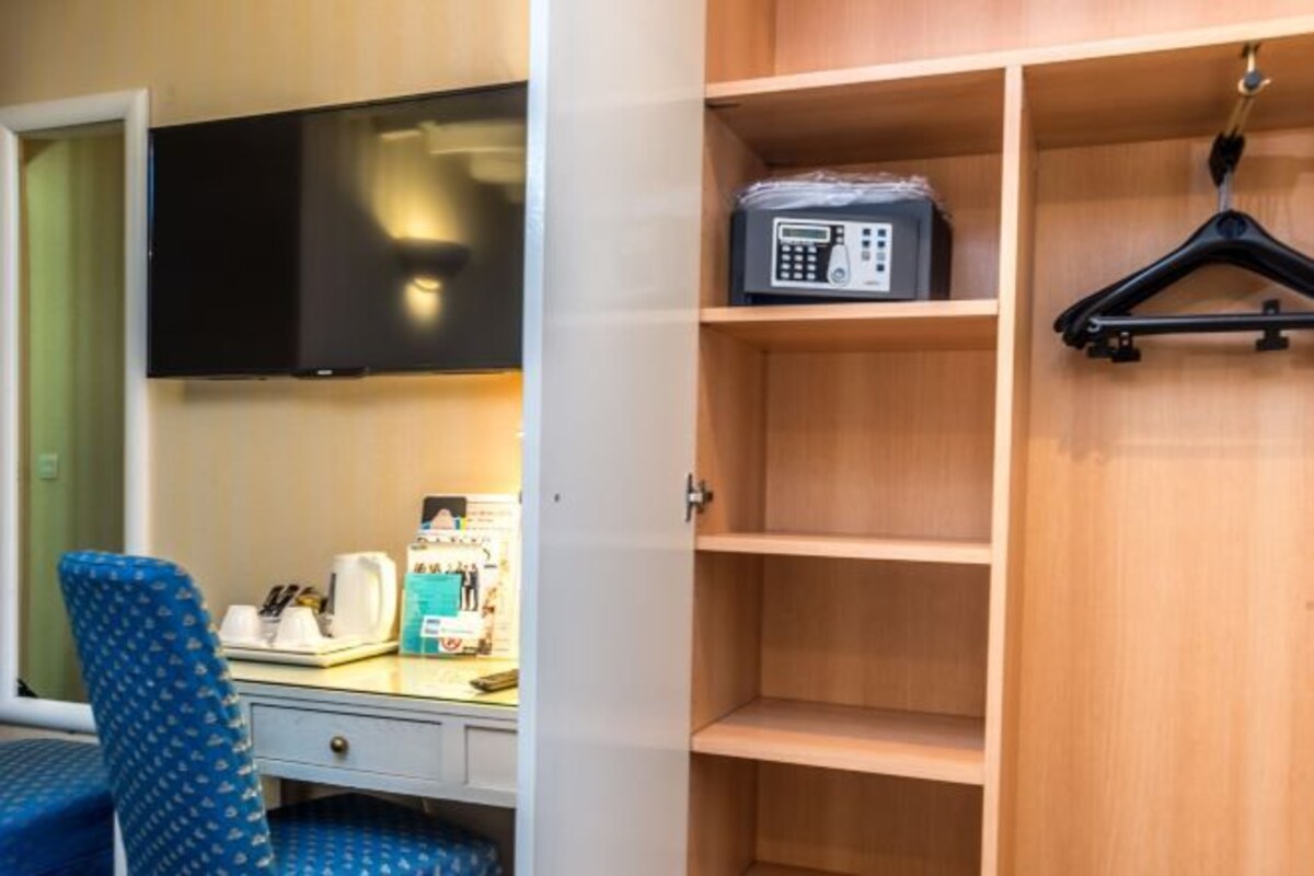 Une chambre d’hôtel dotée d’un téléviseur à écran plat fixé au mur, d’une armoire en bois avec des étagères vides, d’un bureau avec une machine à café et d’une chaise bleue à motifs.