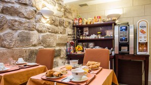 A breakfast setting with tables set for two, a stone wall, a shelf with breakfast items, and a beverage dispenser. - Austin's Arts et Métiers Hôtel (Paris)