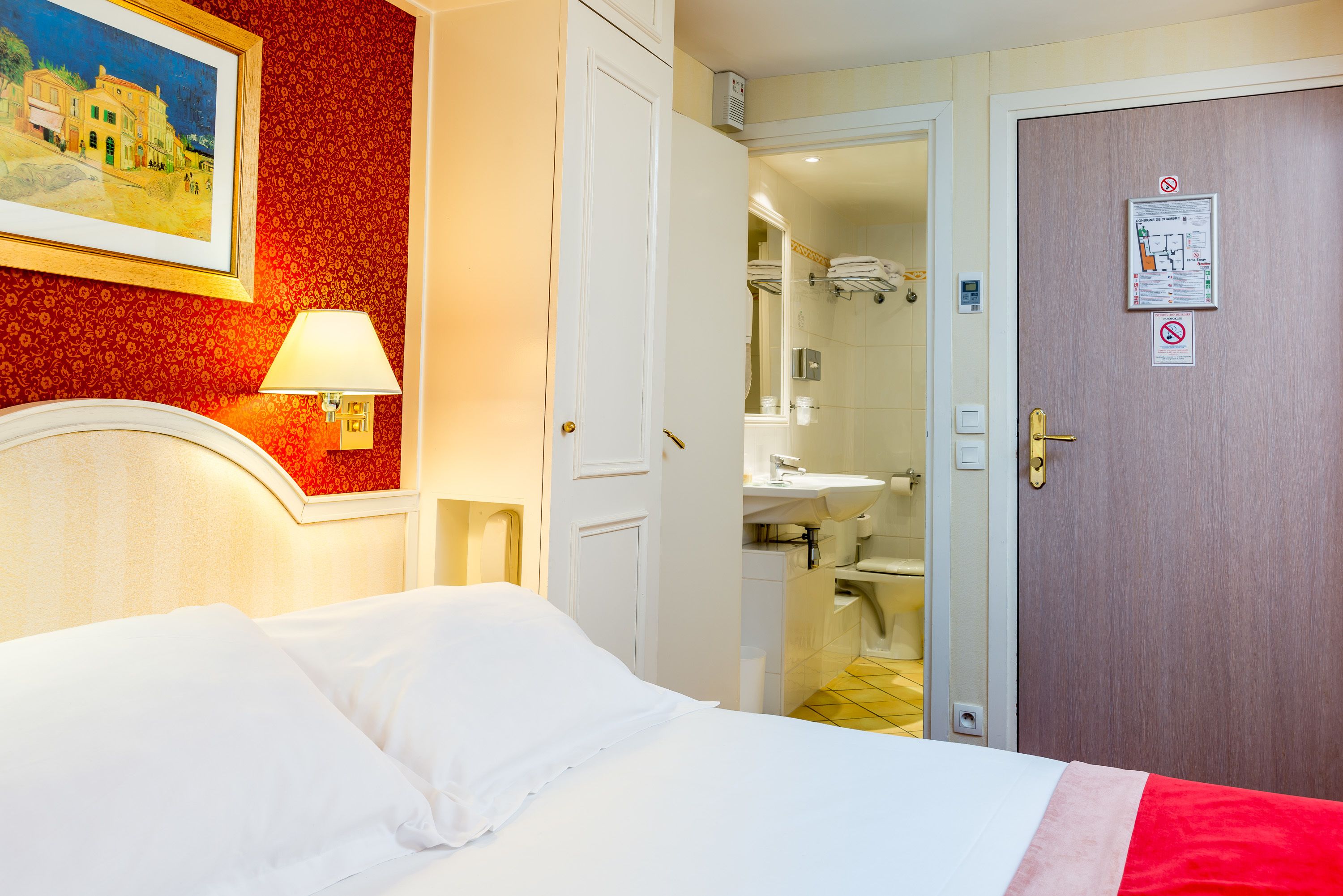 Une chambre d’hôtel avec un lit, un tableau au mur, une lampe et une porte donnant sur une salle de bain.