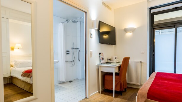 Habitación de hotel con cama, escritorio, silla, televisor y zona de ducha.