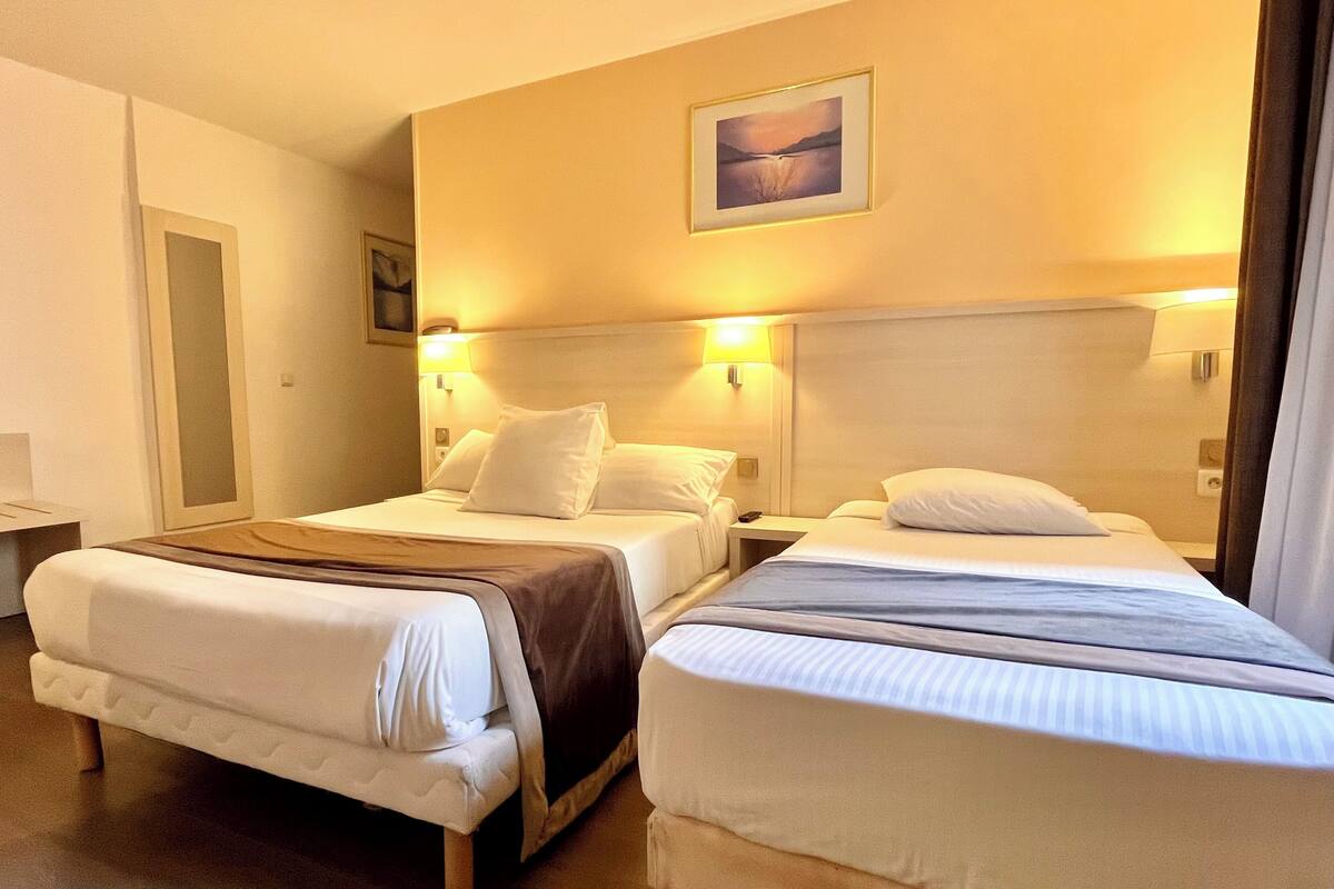 A hotel room with two beds, each with white linens and a brown throw blanket. There are wall-mounted lamps on either side of the beds.