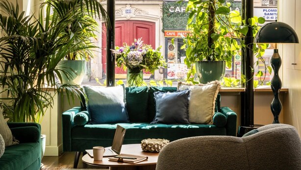 Una sala de estar acogedora con un sofá verde, una mesita de centro y una computadora portátil. Hay plantas en macetas y se ve un café a través de la ventana.