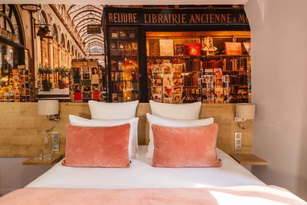 A hotel room with a large bed, two white pillows, and a pink accent pillow. In the background, there is a view of a bookstore with a sign that reads "LIBRAIRIE ANCIENNE & MODERNE".
