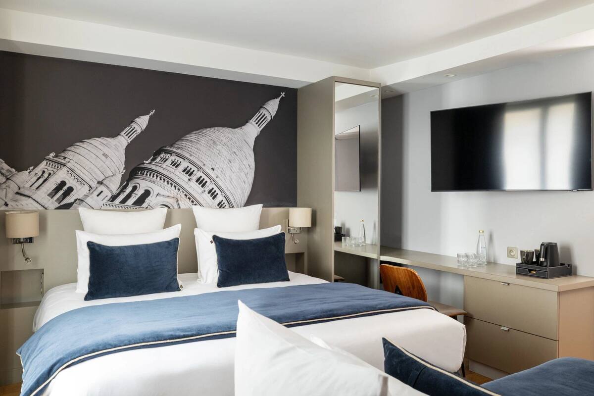 A modern hotel room with a large bed, a flat-screen TV, a desk with a mirror, and a wall mural of a famous building.