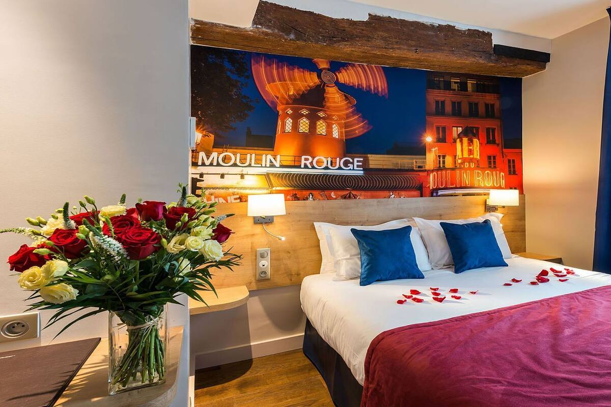 A hotel room with a bed, a nightstand, a vase of flowers, and a wall with a cityscape mural.