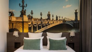 Habitación de hotel con una cama grande, dos almohadas verdes, una colcha blanca y un mural de un puente en la pared.