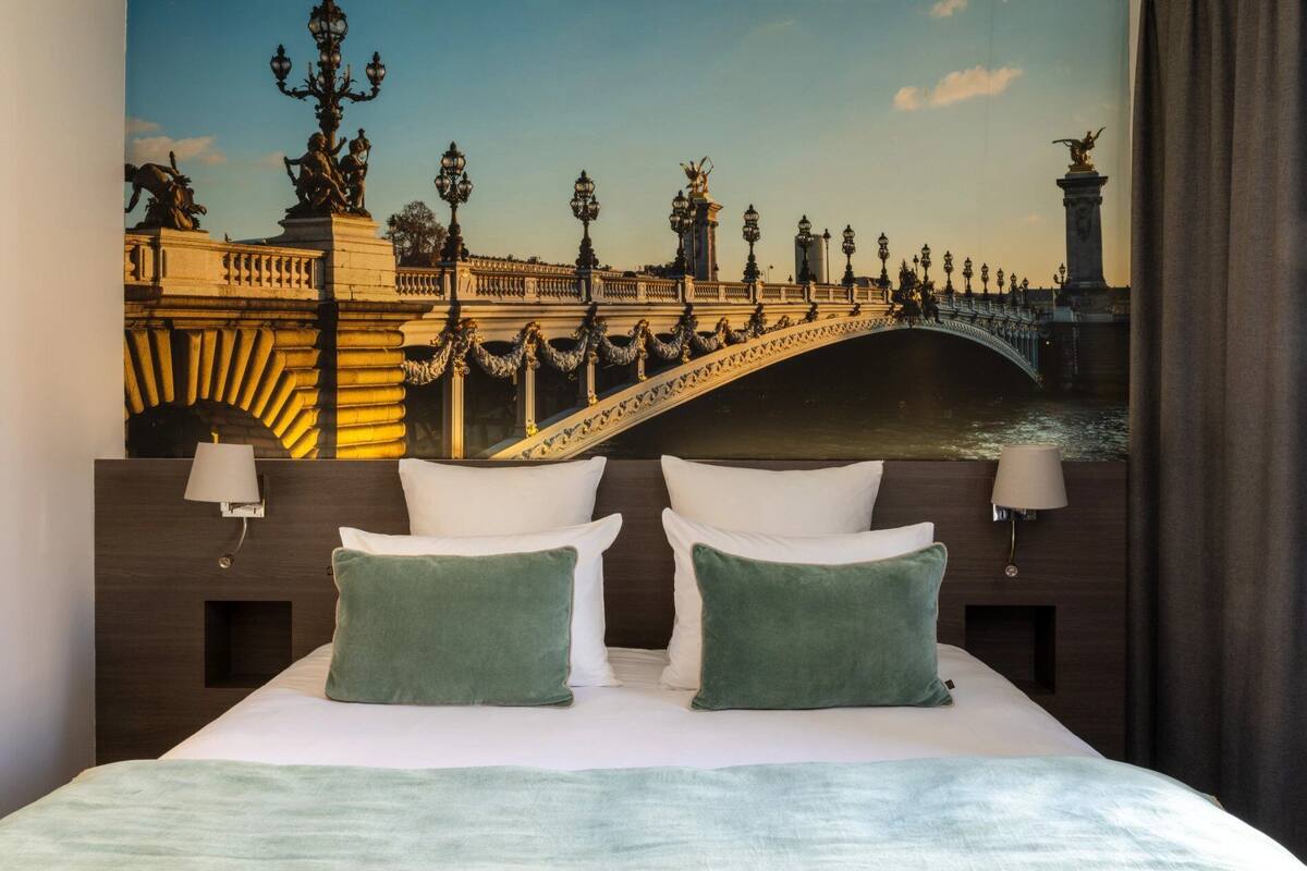 A hotel room with a bed, two pillows, a nightstand, a lamp, and a mural of a bridge.