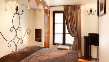A bedroom with a large bed, a window with curtains, a small shelf with decorative items, and a television on a stand.